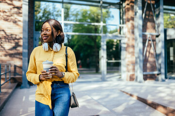 Successful charming afro american woman in yellow blouse headphones, reusable coffee cup waiting outdoor. Eco friendly coffee tea cup mug concept