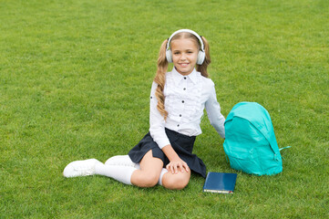 Happy schoolchild in uniform listen to music in headphones sitting on green grass, school holidays