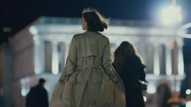 Stylish young woman tourist walking in beautiful square, wearing cozy outfit and coat. City people. Nightlife, travel and urban concept.