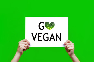 White sheet with a slogan go vegan in female hands isolated on a color green background. A call for...