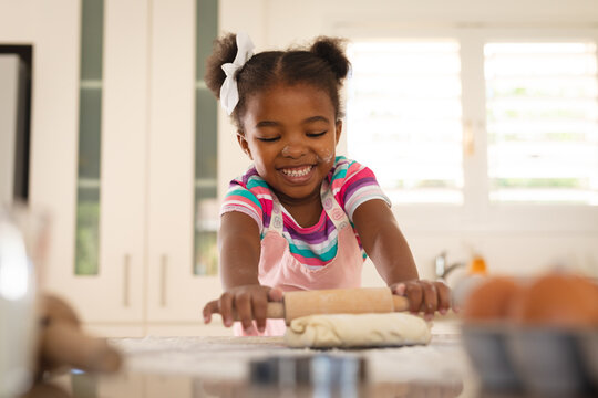 Happy african american daughter leaning on kitchen counter rolling dough with rolling pin