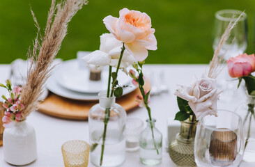 Beautiful multi-colored roses and reeds stand in a glass vase against the background of a wooden table with dishes at a banquet. Photography, concept.