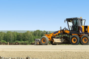 Obraz premium An orange grader is busy with road repairs. Excavator for road works. Road machinery works against the background of the forest.