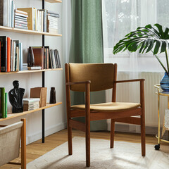Stylish composition of home office interior with design retro chair, home library, plant, window, books, decoration and elegant personal accessories in home decor.