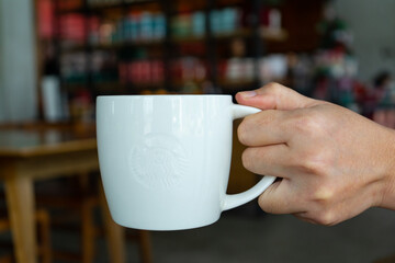 Holding white cup of hot coffee latte or cappuccino on the hand with smoke steam with table and chairs background at the cafe shop restaurant.