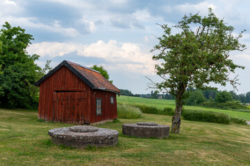 red wooden cottage in country landscape with old millstones in the grass in the foreground