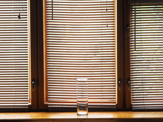 A transparent glass vase on a wooden window sill with closed blinds. Minimalist concept.