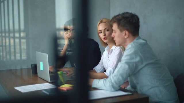 Tracking shot of creative young stratup team of three people discussing business plan while sitting at table in modern office. Male and female business colleagues having discussion looking at laptop.