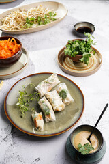 Several spring rolls with chicken, carrot and bamboo sprouts in rice paper on green plate, sesame sauce and coriander on concrete surface