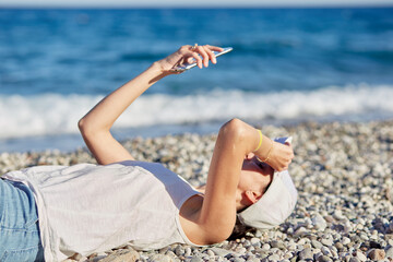Fototapeta na wymiar Young woman is lying in a hat with a phone on the beach near the sea is making a selfie. Resting, traveling and relaxing concept. Copy space