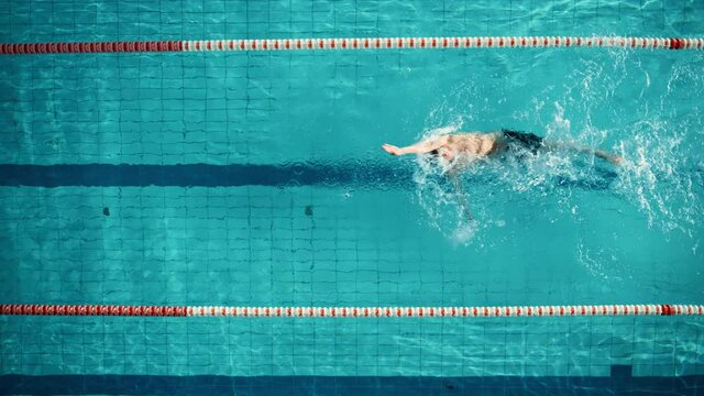 Aerial Top View: Muscular Male Swimmer in Swimming Pool. Professional Athlete Swims in Backstroke Style, Determination in Training to Win Championship. Cinematic Slow Motion, Stylish Colors