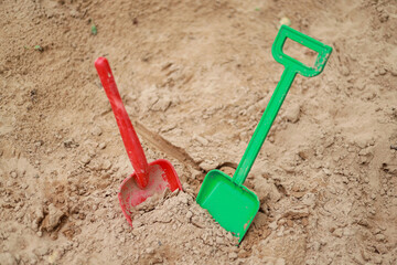Red and green kids toddlers plastic play shovels laying on a sandbox sand background.