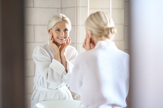 Gorgeous mid age adult 50 years old blonde woman standing in bathroom wearing bathrobe touching face, looking at reflection in mirror smiling doing morning beauty routine. Antiage skin care concept.