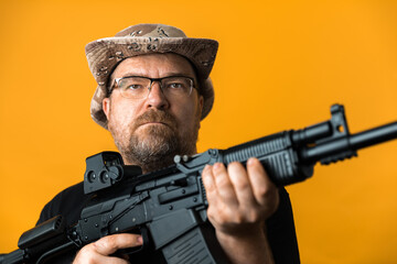 Fototapeta premium Middle aged man with rifle in black t-shirt and hat against yellow background.
