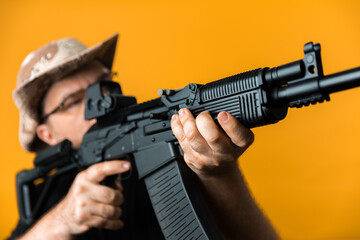 Middle aged man with rifle in black t-shirt and hat against yellow background.