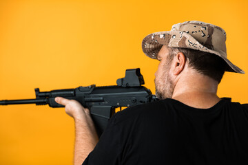 Middle aged man with rifle in black t-shirt and hat against yellow background.