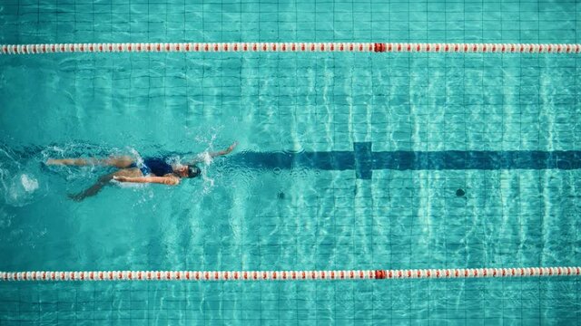 Aerial Top View: Athletic Female Swimmer in Swimming Pool. Professional Athlete Swims in Backstroke Style, Determination in Training to Win Championship. Cinematic Slow Motion, Stylish Colors