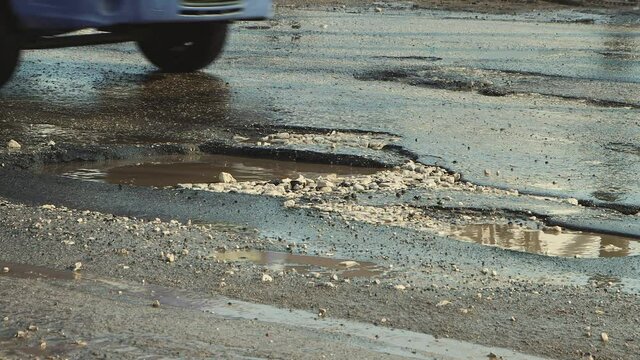 Cars drive in close-up on a bad, bumpy road with holes. Large potholes after rain, damaged road infrastructure. Asphalt in violation of technology. Taxpayer money is wasted and corruption is rampant.