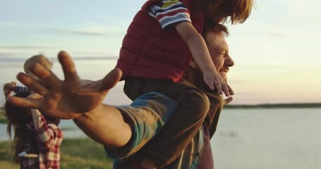 Little cute boys sit on the necks of their mother and father and whirl with them. They are on a hike, you can see a field, a reservoir and a wide sky. The family is having fun