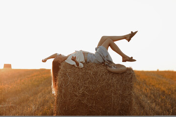 Young woman with long hair, wearing jeans skirt, light shirt is lying on straw bale in field in...