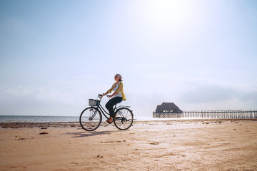 Fototapeta na wymiar Young woman dressed light summer clothes riding old vintage bicycle with front basket on the lonely low tide ocean white sand coast on Kiwengwa beach on Zanzibar island, Tanzania.