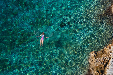 Young female floating on the back and relaxing on the warm turquoise Adriatic sea waves with rocky coastline. Carefree people vacation time concept aerial top view image.