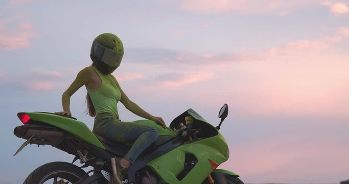 A sexy and athletic young woman in a green jersey sits on a racing motorcycle. She has a protective helmet on her head, she is stained with paint. Behind her, there is a great view of the colorful sky