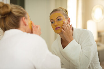 Fototapeta premium Caucasian woman in bathroom wearing bathrobe, looking in mirror and applying face mask