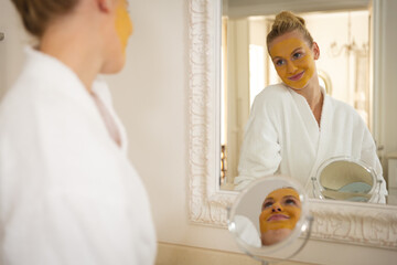 Caucasian woman in bathroom wearing bathrobe wearing beauty treatment face mask, smiling in mirror