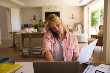 Caucasian woman sitting at table working in living room, using laptop and talking on smartphone