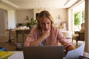 Caucasian woman sitting at table working in living room using laptop and reading paperwork