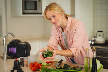 Smiling caucasian woman in kitchen holding vegetables and looking at camera, making cooking vlog