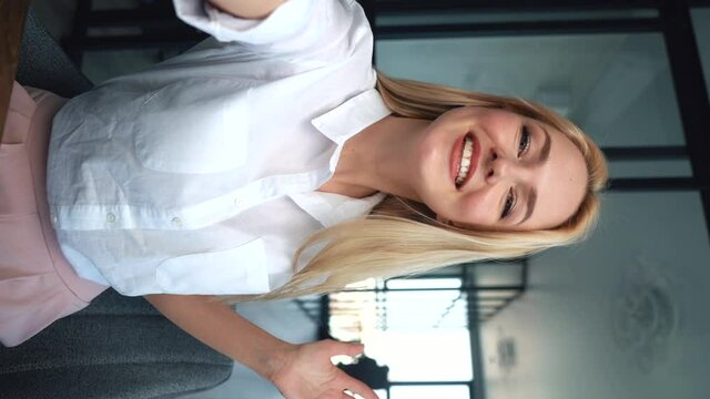 Vertical orientation video of attractive female blogger doing video chat from apartment. Close-up portrait of smiling young blonde woman making selfie or video call while at office in workplace.