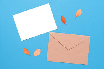 Blank greeting card mockup, brown envelope and autumn leaves on a blue pastel paper background. Autumn stationery wedding template. Top view, flat lay.