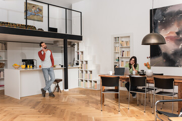 Man talking on cellphone near smiling girlfriend working on laptop at home