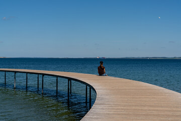 slim woman sits alone on a circular boardwalk leading out and over a calm blue ocean