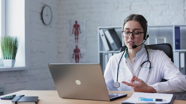 Shot of focused young woman doctor in lab coat, glasses and wireless headset talking on-online video call on laptop while sitting at desk in clinic. Close-up. Health concept. Female doctor speaking.