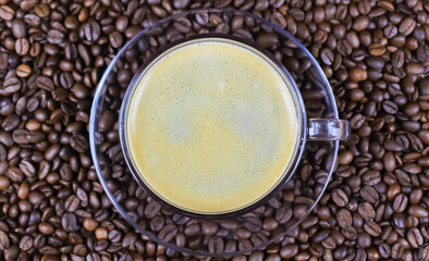 Top view on transparent glass cup, saucer and handle with black coffee whitecap crema, blurred roasted coffee beans background (focus on foam head in cup)