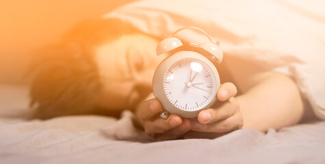 Hands of a young man from under the blankets hold a retro vintage alarm clock in gray. The person...