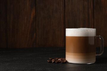 Glass cup of delicious layered coffee and beans on black table against wooden background, space for text