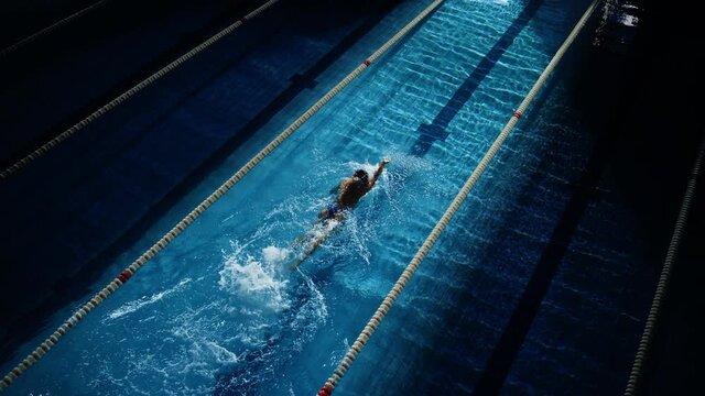Female Swimmer Racing in Swimming Pool. Professional Athlete Overcoming Stress and Hardships in Dark Dramatic Pool, Cinematic Lap Lane Light Showing the Good Way. Aerial Slow Motion Shot