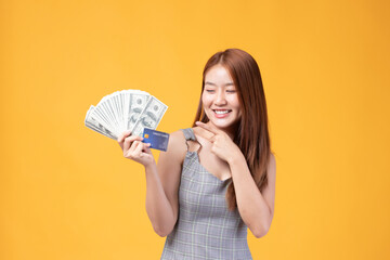 portrait smiling Asian woman in gray dress holding money with credit card isolated on yellow...