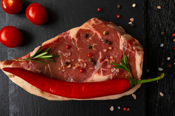 Raw piece of ribeye beef steak on slate board on a wooden table hot pepper smile, spice, himalayan salt, tomato. Fresh food. Everything for a barbecue. Macro