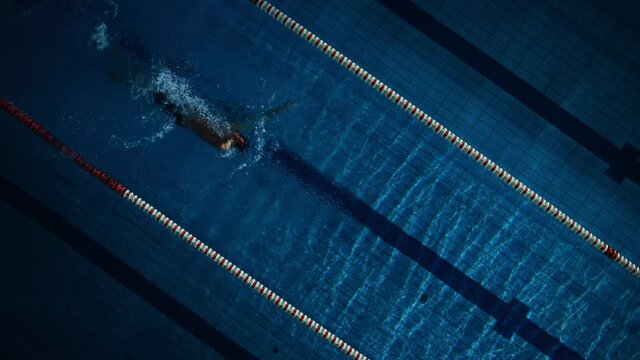 Aerial Top View: Muscular Male Swimmer in Swimming Pool. Professional Athlete Swims in Backstroke Style, Determination to Win Championship. Dark Dramatic Colors, Cinematic Light, Artistic Slow Motion