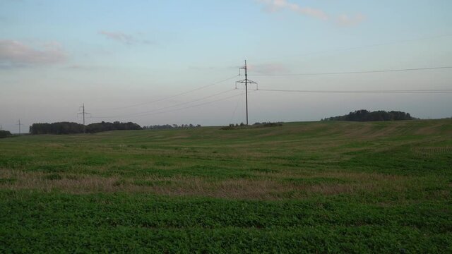 A grass-covered agricultural land scene in the evening with poles of the high voltage line. Countryside panoramic landscape with a little bit cloudy, blue sky. Farmland field set aside.