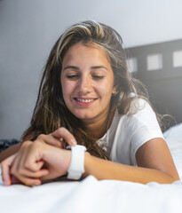 young hispanic woman lying in bed in pajamas smiling using her smart watch