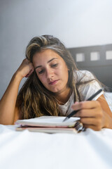 young left-handed hispanic woman lying in bed in her pajamas writing notes in her personal diary