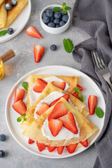 Crepes or thin pancakes with cream cheese, fresh strawberries and honey on a white plate on a gray concrete background. Copy space.