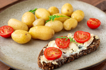 Fototapeta na wymiar Boiled young potatoes and cereal bread with blue cheese and tomatoes on a plate on a wooden background. Vegetarian food.
