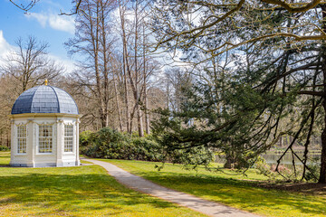 Field with a dirt trail, a Tea dome or theekoepel or Gloriette, green grass, bare trees and a pond, sunny spring day with a blue sky in Paleispark Kroondomein Het Loo in Apeldoorn, Netherlands